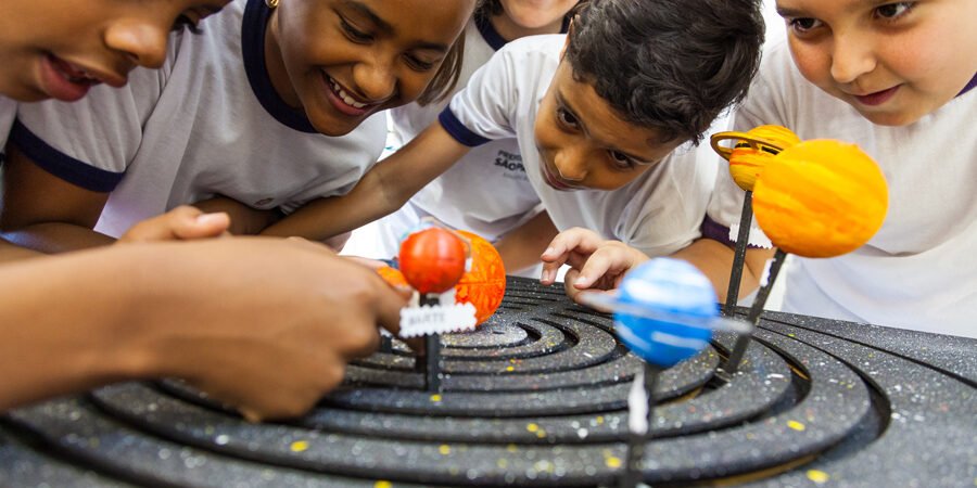 Fotografia de cinco crianças sorridentes, com uniformes escolares brancos, reunidas com curiosidade ao redor de uma maquete tridimensional do sistema solar. Elas estão debruçadas observando os planetas coloridos em miniatura, que estão fixados em órbitas sobre uma base preta circular simulando o espaço.