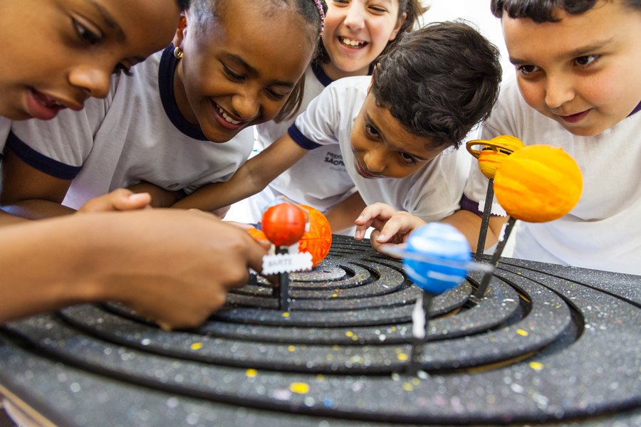Fotografia de cinco crianças sorridentes, com uniformes escolares brancos, reunidas com curiosidade ao redor de uma maquete tridimensional do sistema solar. Elas estão debruçadas observando os planetas coloridos em miniatura, que estão fixados em órbitas sobre uma base preta circular simulando o espaço.