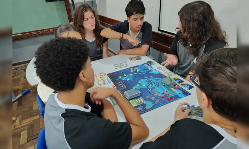 Fotografia de ângulo alto de seis adolescentes ao redor de uma mesa branca em uma sala de aula, jogando WAR. Dois jovens ao fundo gesticulam enquanto discutem a próxima jogada, cercados por outros concentrados. Cartas e fichas estão sobre a mesa.